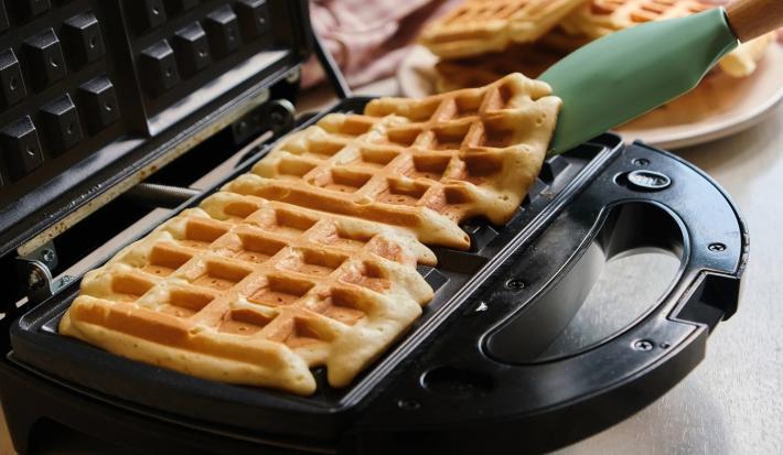 Imagem de uma máquina para waffles com waffles quadrados e uma espátula verde.