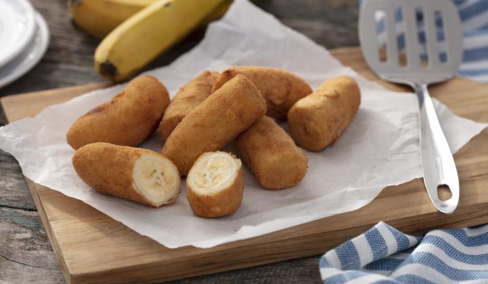 Fotografia em tons de amarelo e azul em uma bancada de madeira clara, um pano azul com listras brancas, uma tábua de madeira com várias bananas à milanesa em cima dela. Ao lado, uma concha de fritura e ao fundo um cacho de bananas.