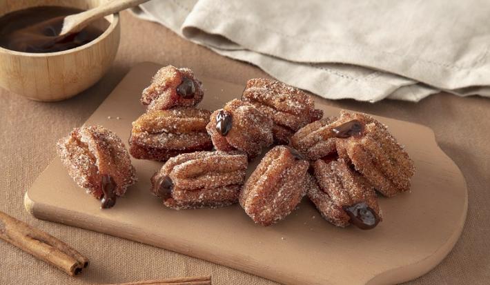 Foto em tons de bege da receita de mini churros recheados com chandelle servida em diversas porções sobre uma tábua de madeira em cima de uma mesa bege com um paninho cinza ao fundo