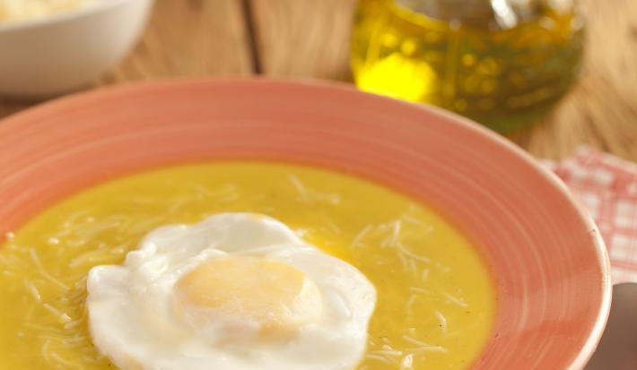 Fotografia em tons de laranja em uma bancada de madeira escura, um pano desenhado laranja, um prato laranja com a sopa portuguesa com ovo pochê dentro dele. Ao fundo, um vidrinho de azeite e um potinho com queijo ralado.