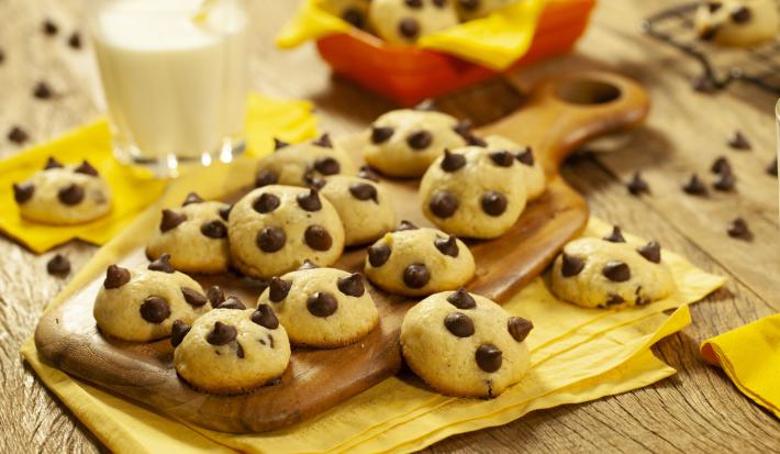 Fotografia em tons de marrom e amarelo de uma bancada de madeira com um paninho amarelo, sobre ela uma tábua de madeira com cookies. Ao fundo um copo de vidro com leite e um recipiente vermelho com um paninho amarelo com cookies.