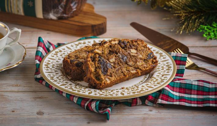 Foto aproximada de uma fatia de bolo de frutas natalino, sobre um prato redondo com bordas decoradas, em uma bancada de madeira onde há decorações de natal, alguns talheres de metal e uma xícara de chá