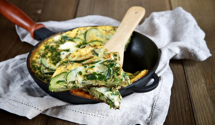 Foto da receita de omelete de abobrinha, servida em uma frigideira preta de ferro, com uma fatia mais próxima, sobre uma colher de pau. A frigideira está sobre um pano bege em uma bancada de madeira.
