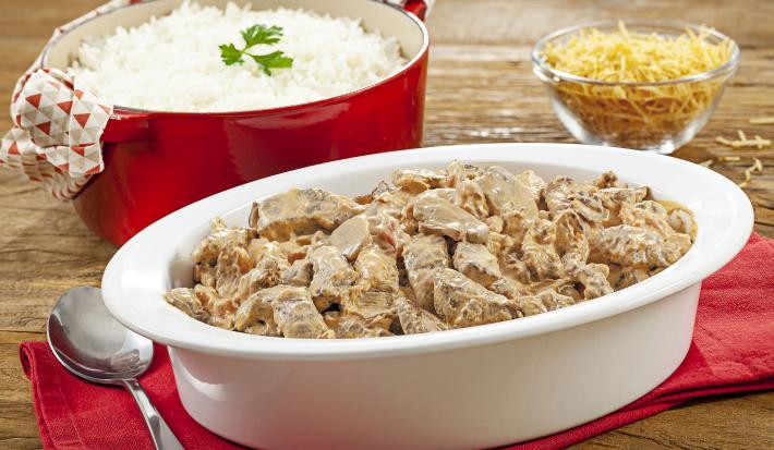 Fotografia em tons de vermelho em uma bancada de madeira com um pano vermelho, uma panela vermelha ao fundo com arroz branco, um potinho de vidro com batata palha ao lado e ao centro, um refratário oval branco com o strogonoff de carne dentro dele.