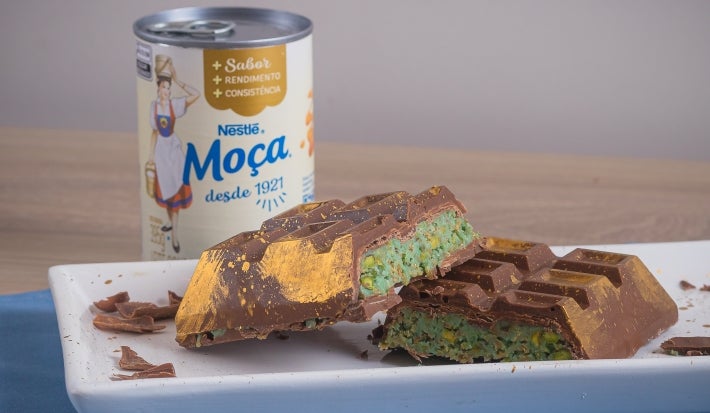 Barra de Dubai, receita de uma barra de chocolate recheada com um creme de pistache e macarrão, decorada com pó dourado e servida em um prato branco ao lado de uma lata de Leite Moça
