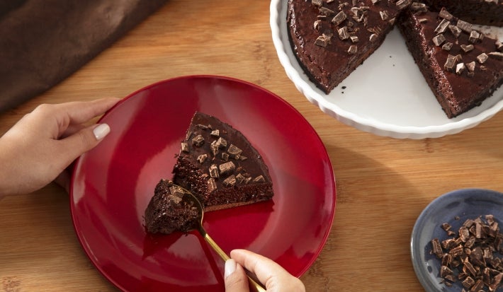 Fotografia de uma mão feminina segurando um prato vermelho raso, sobre uma mesa de madeira. No prato tem uma fatia de torta mousse de nescau extra cacau, ao lado tem um apoio branco com o restante da sobremesa.