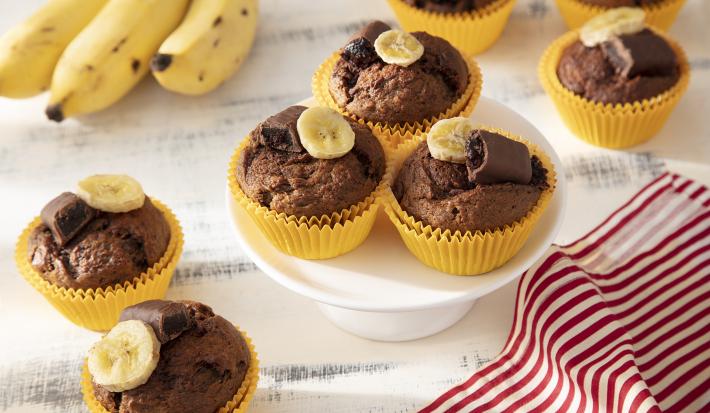 Foto da receita de Muffin de Banana com Chocolate Caribe. Observa-se 8 muffins em forminhas amarelas com uma rodela de banana e metade de um bombom no topo.