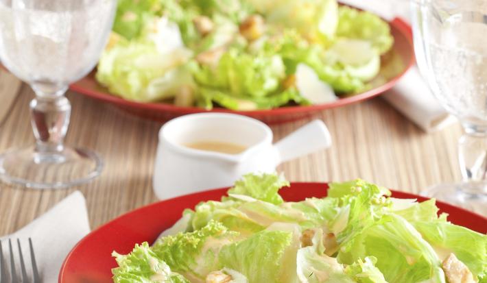 Fotografia em tons de vermelho e verde em uma bancada de madeira clara com um prato vermelho ao centro e a salada caesar com croutons em cima dele.