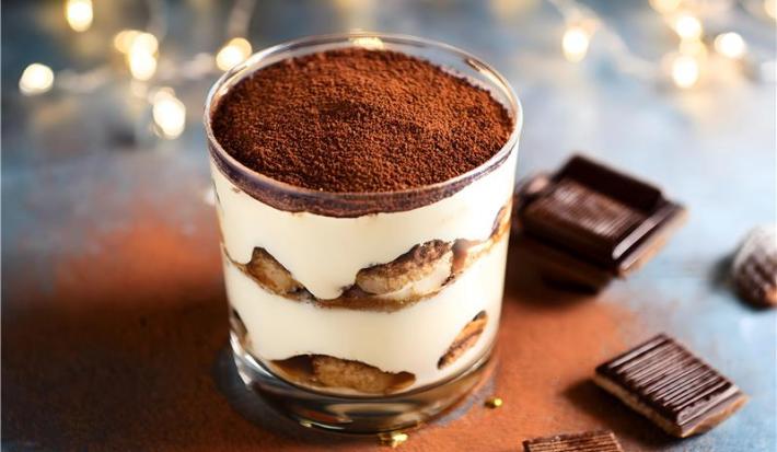 Fotografia de um copo de vidro pequeno com tiramisú. O copo está sobre uma mesa escura com cacau em pó polvilhado e alguns pedaços de chocolate.