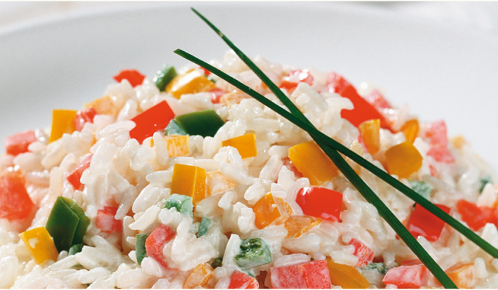 Fotografia em tons de branco e multicoloridos, porcelana branca com porção de arroz com legumes picados e decorados com duas folhas de cebolinha-verde, dois guardanapos brancos ao lado, tudo sobre bancada branca.