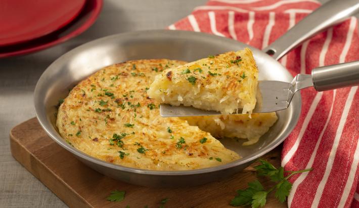 Fotografia em tons de marrom, vermelho e cinza de uma tabua de madeira, uma frigideira de inox com a batata rosti e uma espátula retirando uma fatia. Ao lado um paninho listrado vermelho e branco, ao fundo dois pratos vermelhos.