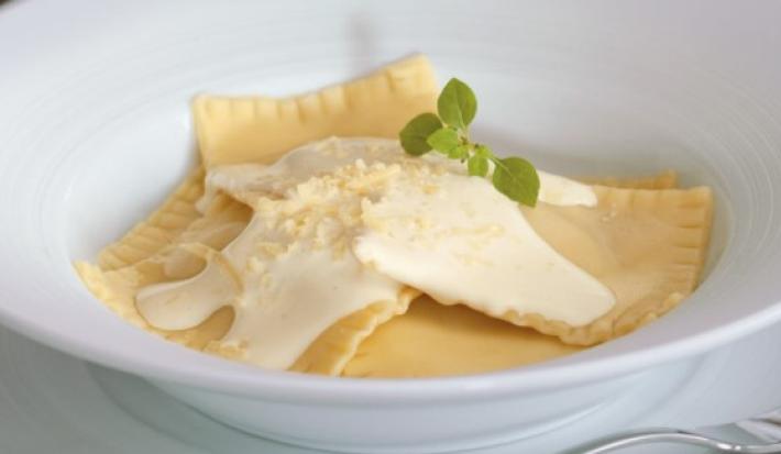Fotografia em tons de amarelo em um fundo branco com um prato redondo grande, largo e branco com ravioli de abóbora com gorgonzola e molho branco em cima.