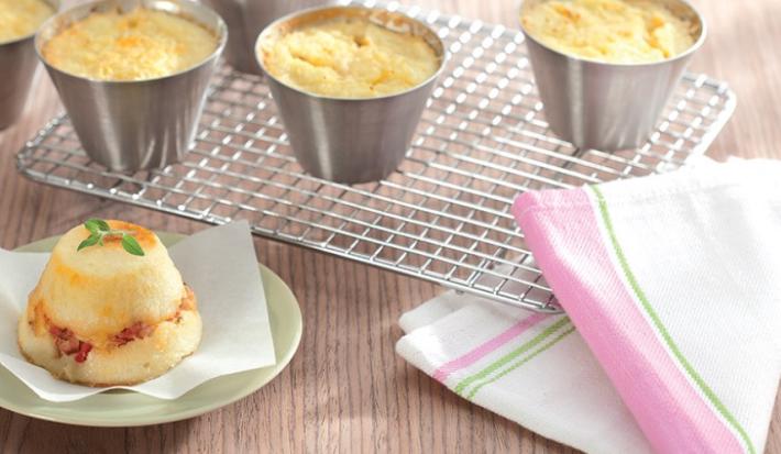 contém 4 potinhos redondos de alumínio com a massa dentro todos por cima de uma grade. Um prato redondo verde com o Muffin e ao lado um pano em tons de branco, rosa e verde.