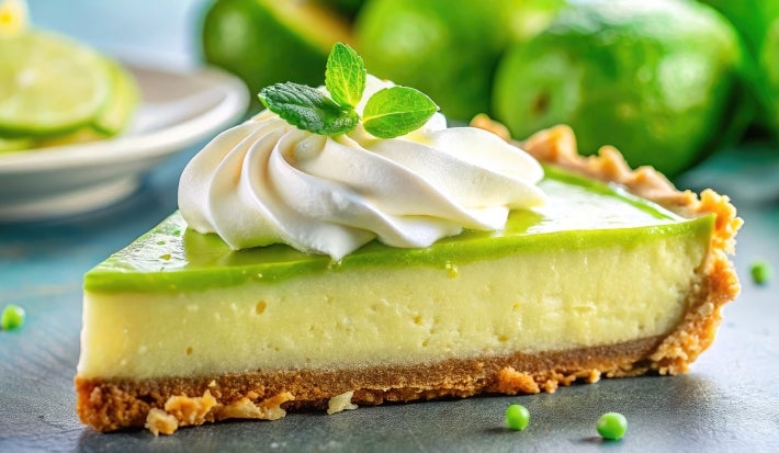 Fotografia em tons claros de uma fatia de torta de limão com gelatina de limão e um pouco de chantilly por cima. Ao fundo tem limões inteiros e um prato de vidro branco e raso com fatias de limão.