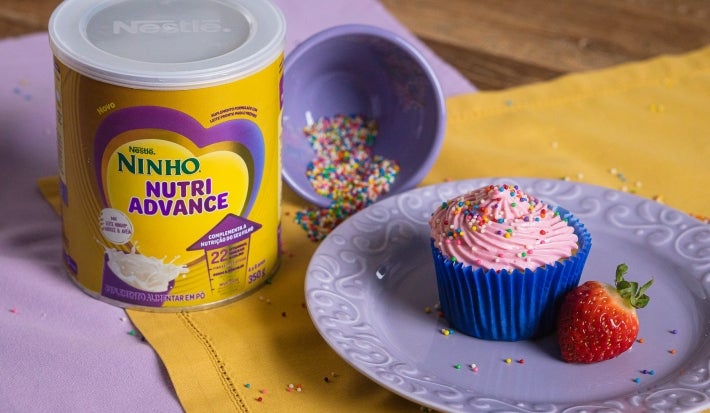 Fotografia de dois panos de mesa, roxo e amarelo, um sobre o outro em uma mesa de madeira. Sobre eles tem uma lata de NINHO Nutri Advance ao lado de um prato médio roxo com um cupcake e um morango inteiro, ao lado tem um potinho roxo com granulados.