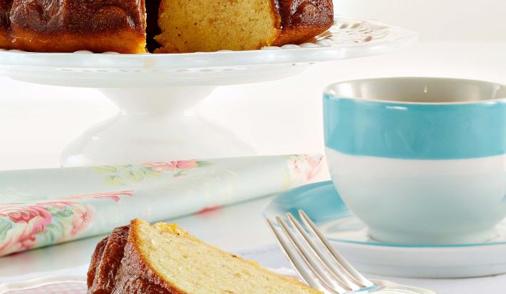 Fotografia em tons de azul em uma mesa branca com uma toalha branca, um prato de sobremesa com uma fatia de bolo de maracujá. Ao fundo, um suporte para bolo com o bolo inteiro e ao lado uma xícara com listra azul e chá dentro dela.