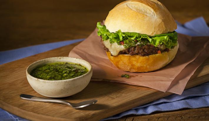 Fotografia em tons de azul em uma bancada de madeira, com uma tábua de madeira, um guardanapo marrom, um hambúrguer caseiro em cima dele. Ao lado, um potinho com molho chimichurri.