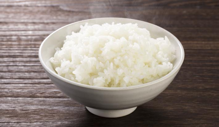 Fotografia em tons de marrom e branco de uma bancada de madeira vista de cima, um recipiente branco redondo contém arroz branco dentro.