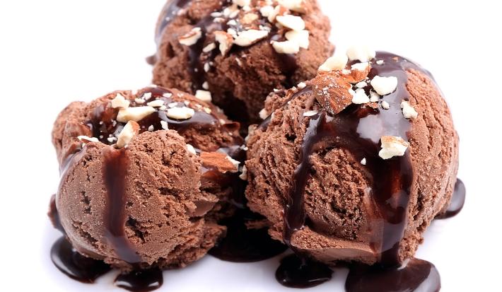 Fotografia de três bolas de sorvete de chocolate com cobertura de chocolate e farofa doce. A sobremesa está em um fundo branco.