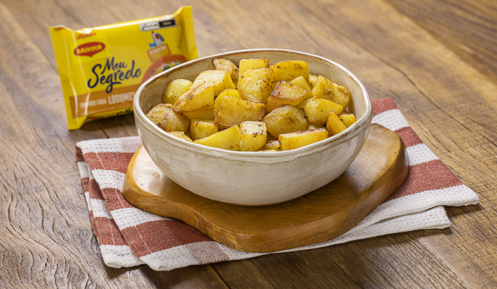Fotografia em tons de amarelo e marrom de uma bancada de madeira com um pano retorcido amarelo, uma travessa branca oval com vários cubos de batatas dentro e salsinha polvilhada.