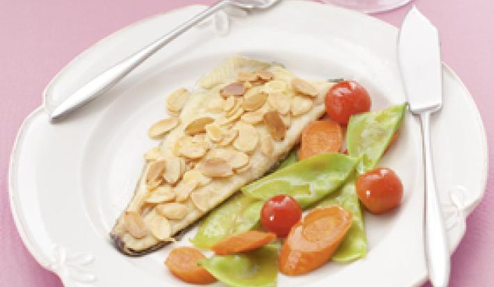 Fotografia em tons de rosa em uma mesa de madeira rosa com um prato branco raso e um filé de truta com amêndoas laminadas e salada dentro dele.