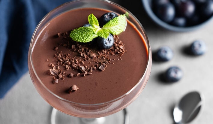 Fotografia de uma taça de vidro com a sobremesa de modo individual, finalizada com ganache, raspas de laranja, hortelã e mirtilos, para variar.