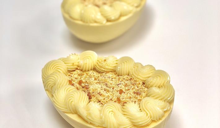 Fotografia em tons de cinza, branco e caramelo de uma bancada branca. Sobre ela, duas cascas de ovos recheadas e decoradas com cobertura.