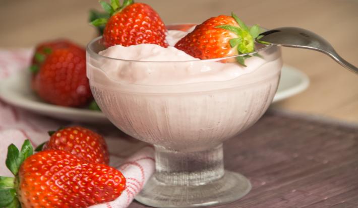 Fotografia de um creme de frutas vermelhas com iogurte, sobre o gelado, dois morangos inteiros e uma colher apoiada no recipiente de vidro em que está o creme. Ao lado, dois morangos, e ao fundo, mais morangos em cima de um prato branco pequeno.