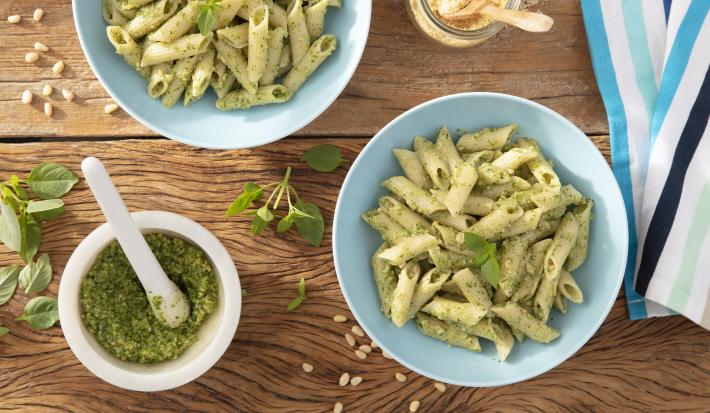 Fotografia em tons de verde em uma bancada de madeira escura, um pano branco listrado com azul, dois pratos azuis com macarrão penne com molho pesto. Ao lado, um potinho branco com o molho pesto dentro dele.