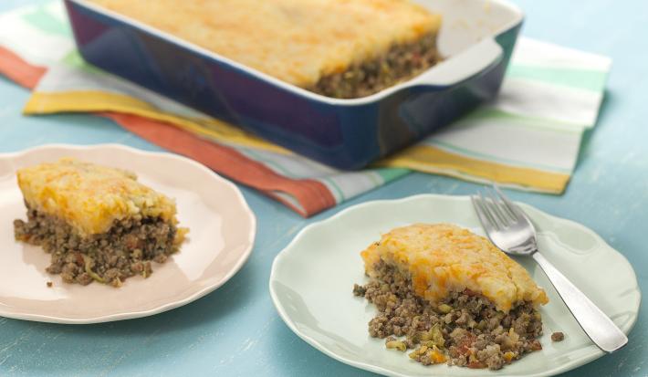 Fotografia em tons de azul em uma bancada de madeira azul, dois pratos redondos rasos com fatias do escondidinho de carne com abobrinha e purê de batata e cenoura em cima de cada um. Ao fundo, um refratário azul escuro com o escondidinho inteiro.