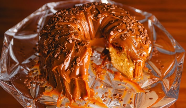 Fotografia em tons de marrom com um bolo de furo no meio ao centro. O bolo possui massa branca, frutas vermelhas e cobertura de brigadeiro de caramelo salgado.