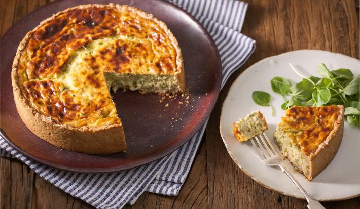 Fotografia em tons de marrom e branco de uma bancada de madeira com um paninho branco e azul, sobre ele um prato redondo vinho com um quiche. Ao lado um prato branco redondo com uma fatia de quiche.