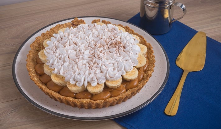 Receita de torta banoffee, servida num prato branco e decorada com chantilly e cacau em pó. A torta está ao lado de uma espátula dourada em uma bancada de madeira com um tecido na cor azul escura