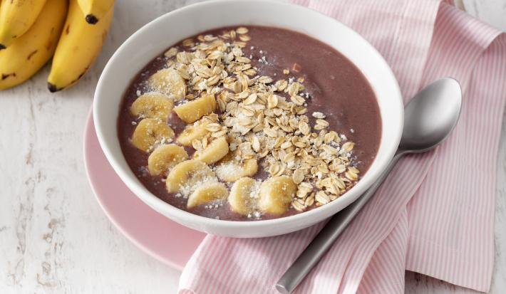 Fotografia em tons de branco e rosa de uma bancada de madeira branca com um paninho rosa, sobre ele um prato raso rosa com um recipiente redondo branco com o açaí. Ao lado uma colher e um cacho de banana.