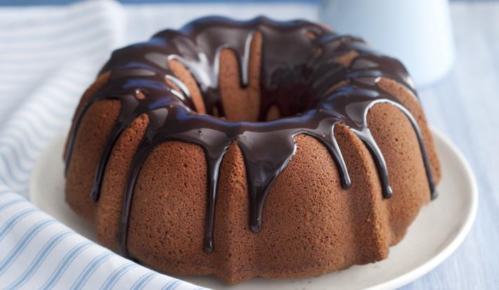Fotografia em tons de azul em uma bancada de madeira clara com um pano branco listrado em azul, ao centro um prato branco raso com o bolo de chocolate, com cobertura de chocolate. Ao lado, um potinho com chocolate e uma jarra azul claro.