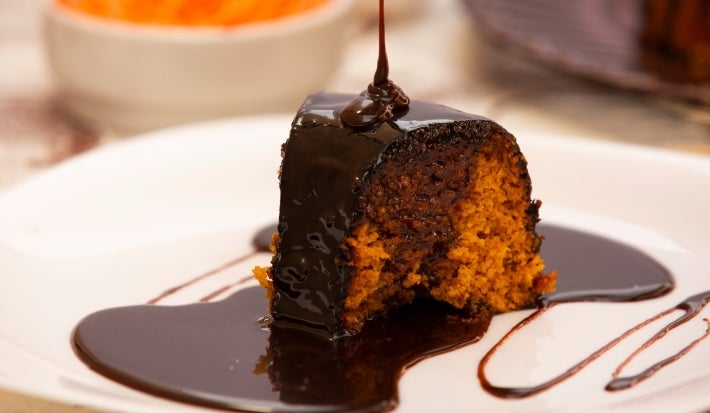 Um pedaço de bolo de cenoura com calda de chocolate escorrendo por cima. A cobertura está sendo servida no momento, o que dá um visual ainda mais apetitoso ao bolo. O interior do bolo parece úmido e bem estruturado.