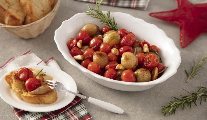 Foto da receita de Confit de Tomates e Cebolas. Observa-se um refratário branco de porcelana com os tomates e as cebolas dentro, acompanhados de alho e alecrim. Ao lado esquerdo, um pratinho com uma torrada acompanhada do confit.