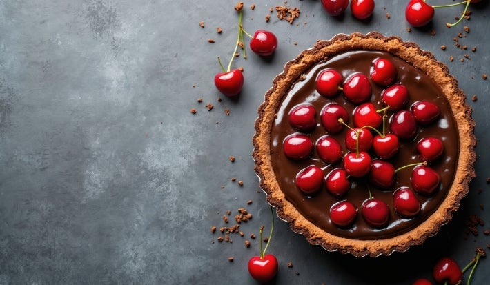 Fotografia vista de cima de uma torta inteira de chocolate e cerejas decorando a torta, que está dentro de uma forma de alumínio sobre uma mesa escura. Ao redor da torta tem cerejas inteiras.