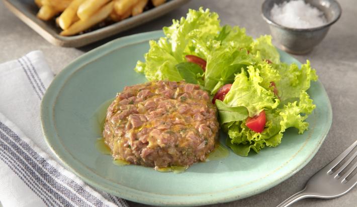 Fotografia em tons de verde em uma bancada de madeira clara, um pano azul, um prato verde claro com o steak tartare acompanhado de salada de alface e tomate cereja. Ao fundo, um recipiente com uma porção de batatas fritas.