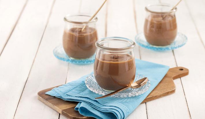 Fotografia em tons de azul em uma bancada de madeira branca, um pano azul turquesa, uma tábua de madeira e um potinho de vidro com a mousse de chocolate dentro dele. Ao fundo, mais potinhos com mousse.