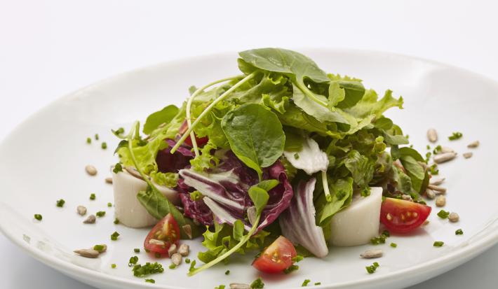 Fotografia de uma salada de agrião, tomate cereja e palmito temperados com vinagrete. A salada está em um prato branco fundo, com algumas sementes e cebolinha.