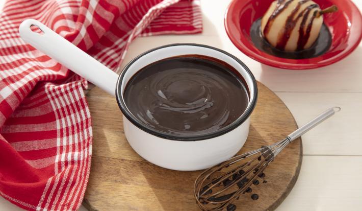 Fotografia em tons de vermelho de uma bancada, ao centro uma panela branca com uma calda de chocolate, em volta um pano vermelho e branco.