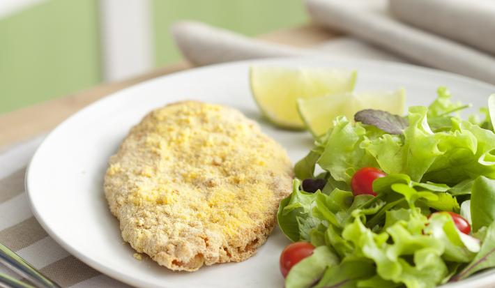 Fotografia em tons de verde em uma bancada de madeira com um pano branco com listras beges e um prato oval branco com o filé de frango empanado e salada verde dentro dele.