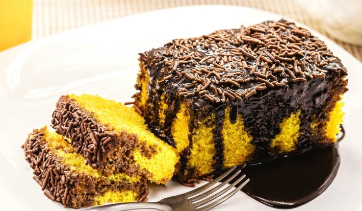 Um pedaço generoso de bolo de cenoura com cobertura de chocolate está em um prato branco. A cobertura é brilhante e farta, com granulado por cima. Ao lado, duas fatias menores destacam a massa amarelo-viva e fofinha, criando contraste com o chocolate