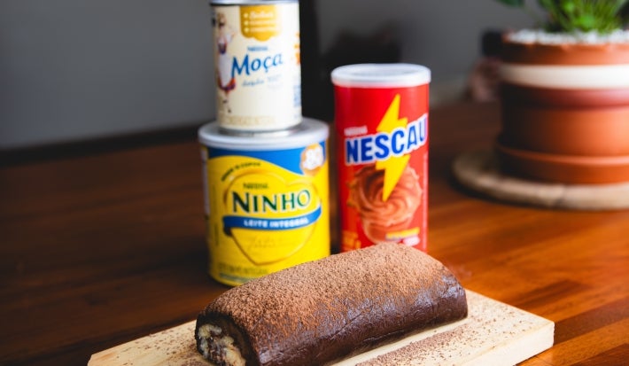 Fotografia em tons terrosos de um rocambole de massa de chocolate recheado com um creme de prestígio. Ao fundo existe uma lata de NESCAU, uma de Leite MOÇA e outra de Leite NINHO em pó Fotografia em tons terrosos de um rocambole de massa de chocolate recheado com um creme de prestígio. Ao fundo existe uma lata de NESCAU, uma de Leite MOÇA e outra de Leite NINHO em pó