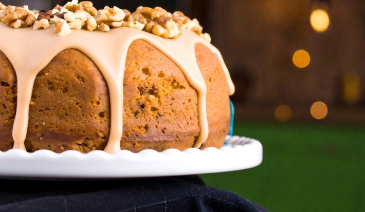 Bolo bundt elegantemente apresentado em um suporte branco. Com uma cobertura cremosa de cor caramelo e decorado com nozes picadas, o bolo exibe uma cor dourado-escura.