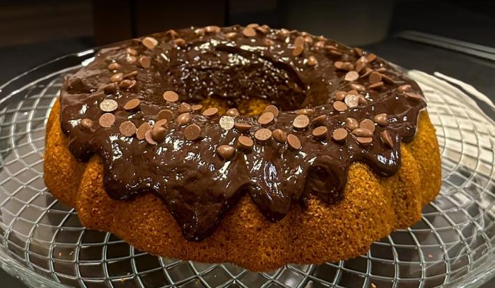 Foto da receita de Bolo de cenoura e especiarias sem glúten. Observa-se um bolo com furo no meio fofinho com uma calda brilhante de chocolate decorado com gotas.
