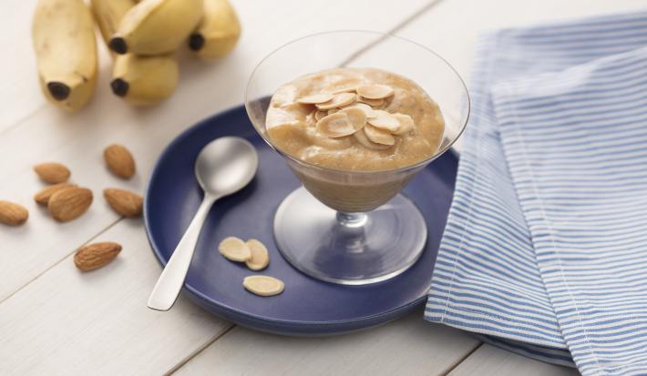 fotografia em tons de branco, azul e marrom de uma bancada vista de cima. Contém uma bandeja redondo e azul com uma taça transparente com creme de banana e amêndoas por cima. Ao lado um pano em tons de azul e branco e do outro uma colher para servir.
