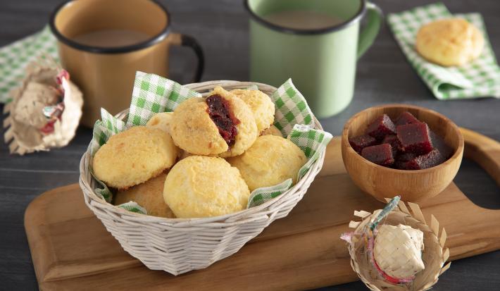 Foto em tons de amarelo e marrom da receita de pão de queijo romeu e julieta servida em diversas porções em uma cestinha sobre uma tábua de madeira com decoração de festa junina e um bowl com goiabada. Ao fundo há duas canecas