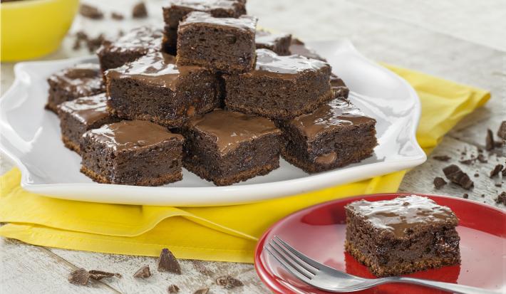 Fotografia em tons de chocolate, branco e amarelo, com prato branco com pedaços de bolo de chocolate sobre guardanapo amarelo, prato vermelho com porção de bolo e um garfo, no alto pote amarelo com chocolate, sobre bancada clara com pedaços de chocolate.